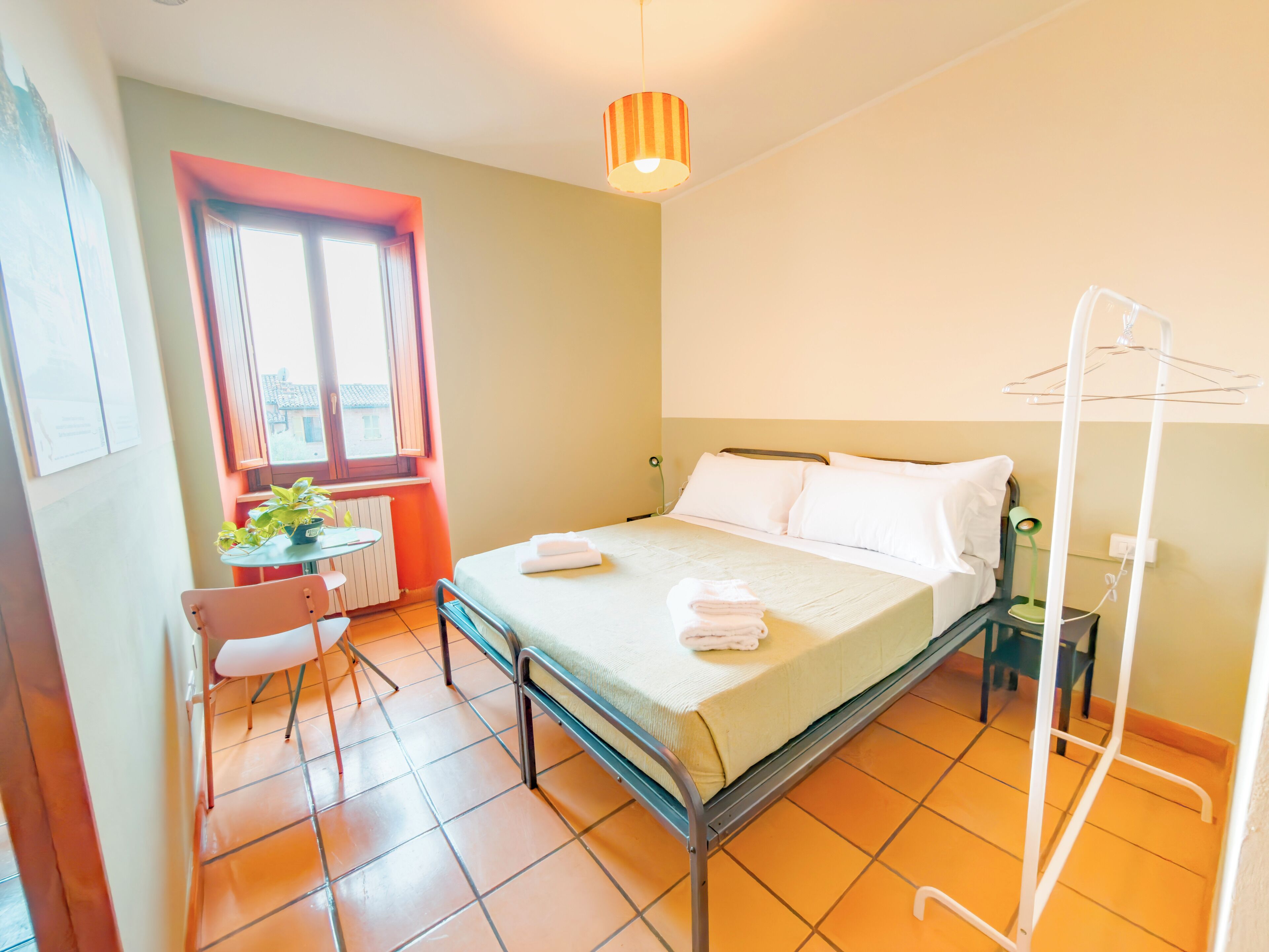A hotel room with a bed, a chair, a table, a radiator, a window, and a hanging clothes rack.