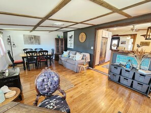 A living room with a dining area, wooden floors, a sofa, a coffee table, a decorative clock, and storage units. - Pony Tales | Near Assateague & Wildlife Refuge (Chincoteague Island)