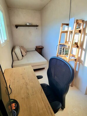 Ein kleiner, kompakter Raum mit einem Einzelbett, einem Holztisch, einem schwarzen Bürostuhl und einem Holzregal.