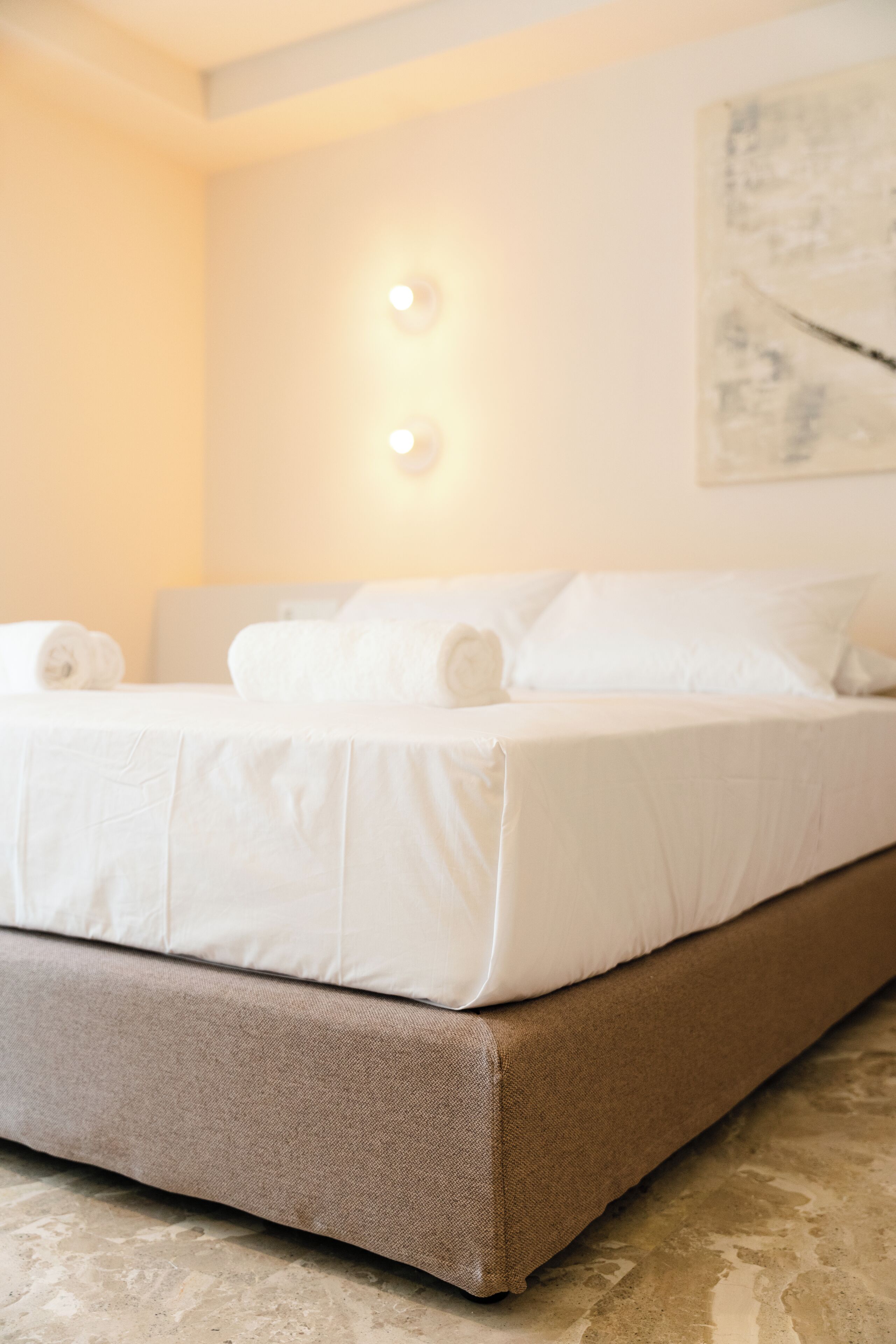 Una cama bien hecha con sábanas blancas y un cabecero marrón en una habitación con pared de color claro y un cuadro.