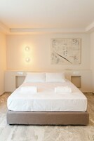 Una cama bien tendida con sábanas blancas y dos toallas, un cuadro abstracto colgado en la pared y dos lámparas de noche.