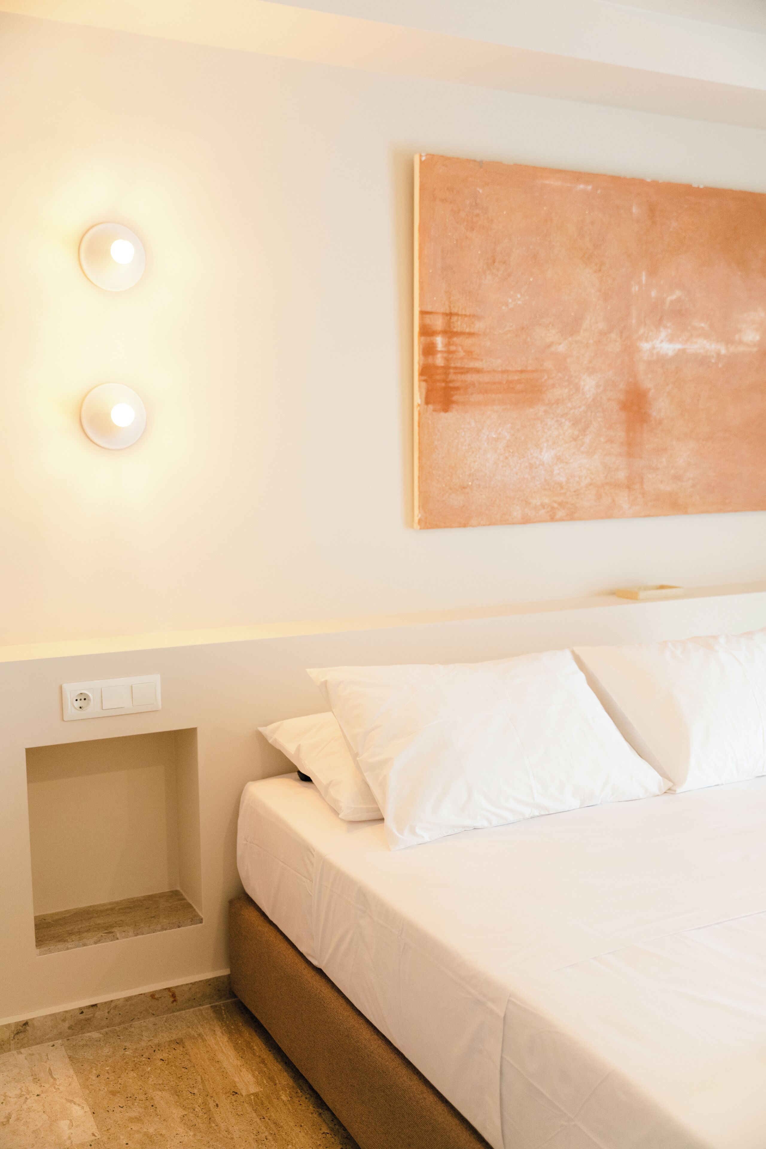 Quarto de hotel com cama, roupa de cama branca, luminária fixada na parede e uma pintura.