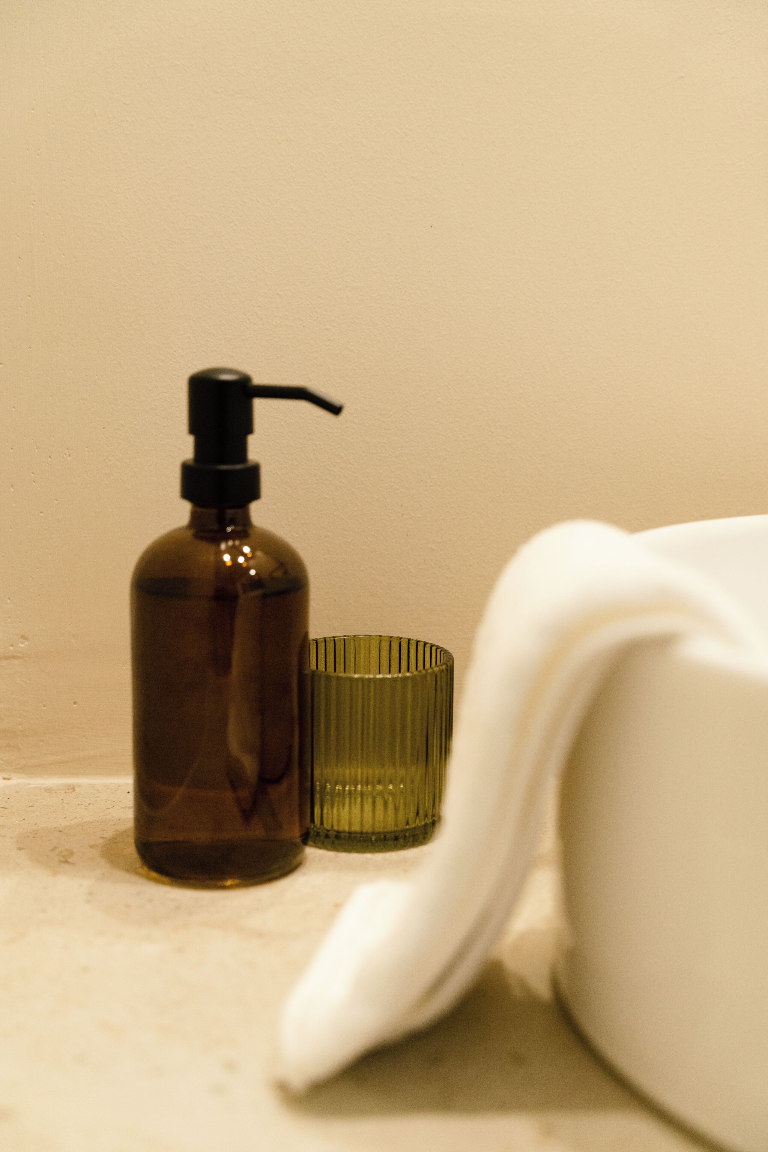 A bathroom with a bottle of liquid soap, a green glass container, and a white towel.
