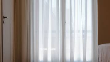 Quarto de hotel com uma janela grande coberta por cortinas finas, uma cama e uma porta.