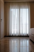 Una habitación de hotel con un ventanal grande cubierto por cortinas de gasa, una cama y una puerta.