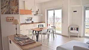 Een moderne keuken met witte kasten, een eettafel met stoelen, een klein zithoekje met een bank en een balkon met een rieten stoel.