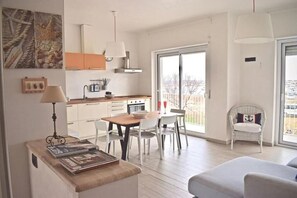 A modern kitchen with white cabinets, a dining table with chairs, a small seating area with a sofa, and a balcony with a wicker chair. - Harbor House II, across from the Marzamemi marina (Marzamemi)