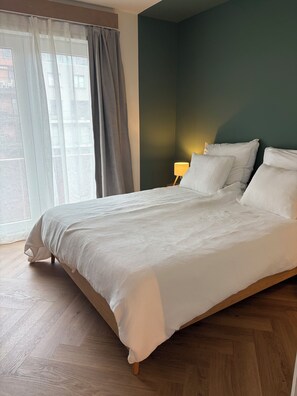 A bedroom with a bed, wooden flooring, a nightstand with a lamp, and a view through a window with curtains. - Apartment for 8 people in Koksijde-Bad—just a stone's throw from the beach (Koksijde)