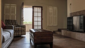 Una sala de estar con sofá, una silla, un reposapiés, televisión y una puerta que da al exterior.