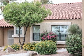 Uma casa com telhado de telhas, uma árvore e plantas floridas no jardim frontal.