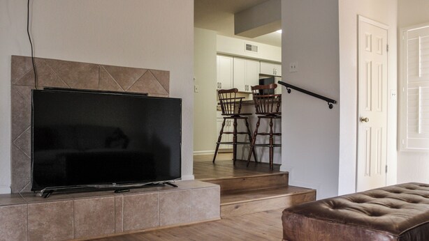 Una sala de estar con un televisor de pantalla plana, un reposapiés de cuero marrón y una escalera que lleva a un comedor.