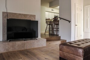 A living room with a flat-screen TV, a brown leather ottoman, and a staircase leading to a dining area.