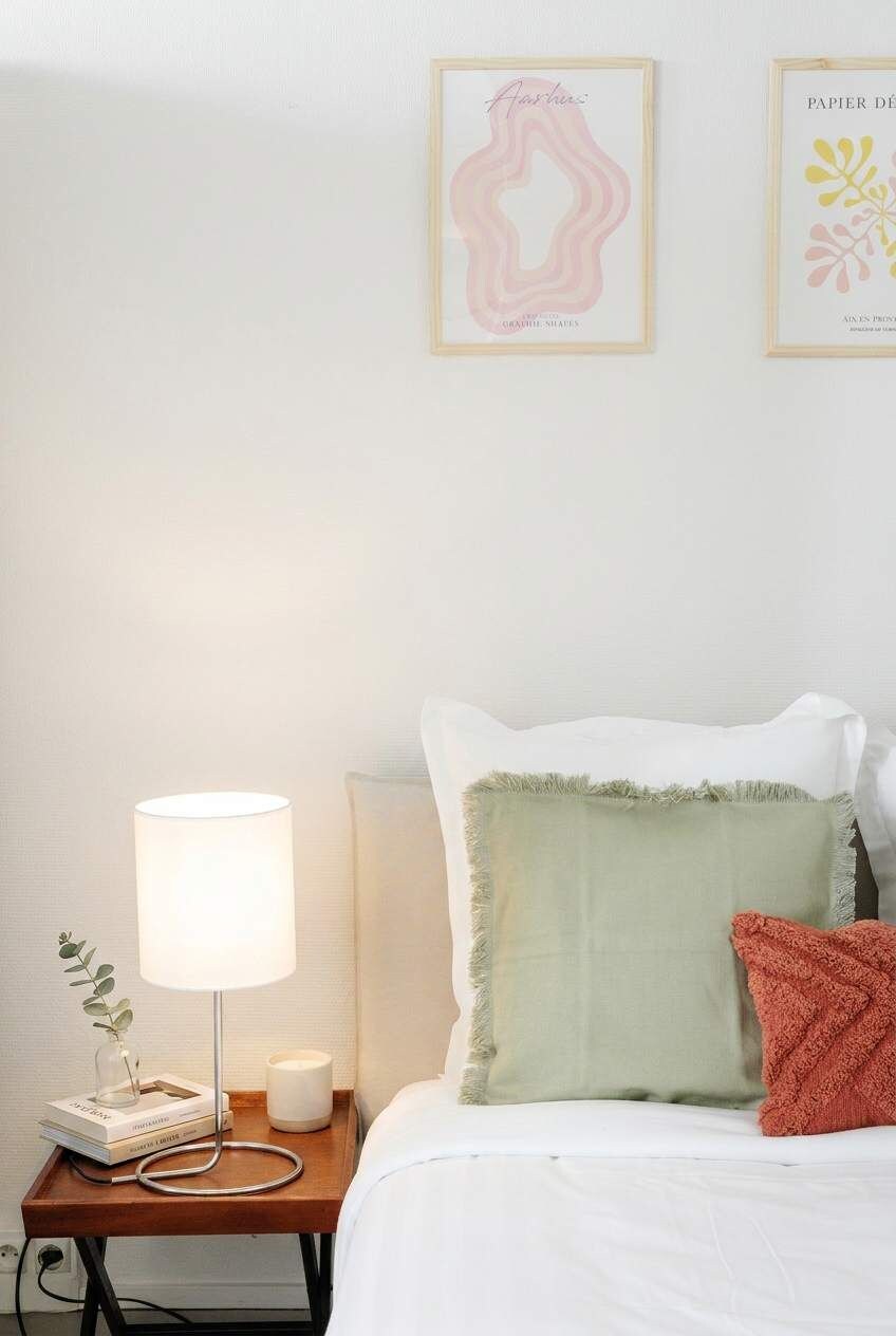 A neatly made bed with pillows, a bedside lamp, a small table with books and a candle, and two framed artworks on the wall.