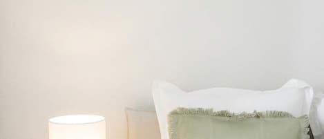 A neatly made bed with pillows, a bedside lamp, a small table with books and a candle, and two framed artworks on the wall.