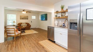 A modern kitchen with stainless steel appliances, a dishwasher, and a refrigerator. There is a dining area with a wooden table and chairs, and a living room with a sofa and a ceiling fan. - The Magnolia on 54th (Columbus)
