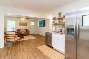 Eine moderne Küche mit Edelstahlgeräten, einem Geschirrspüler und einem Kühlschrank. Es gibt einen Essbereich mit einem Holztisch und Stühlen sowie ein Wohnzimmer mit einer Couch und einem Deckenventilator.