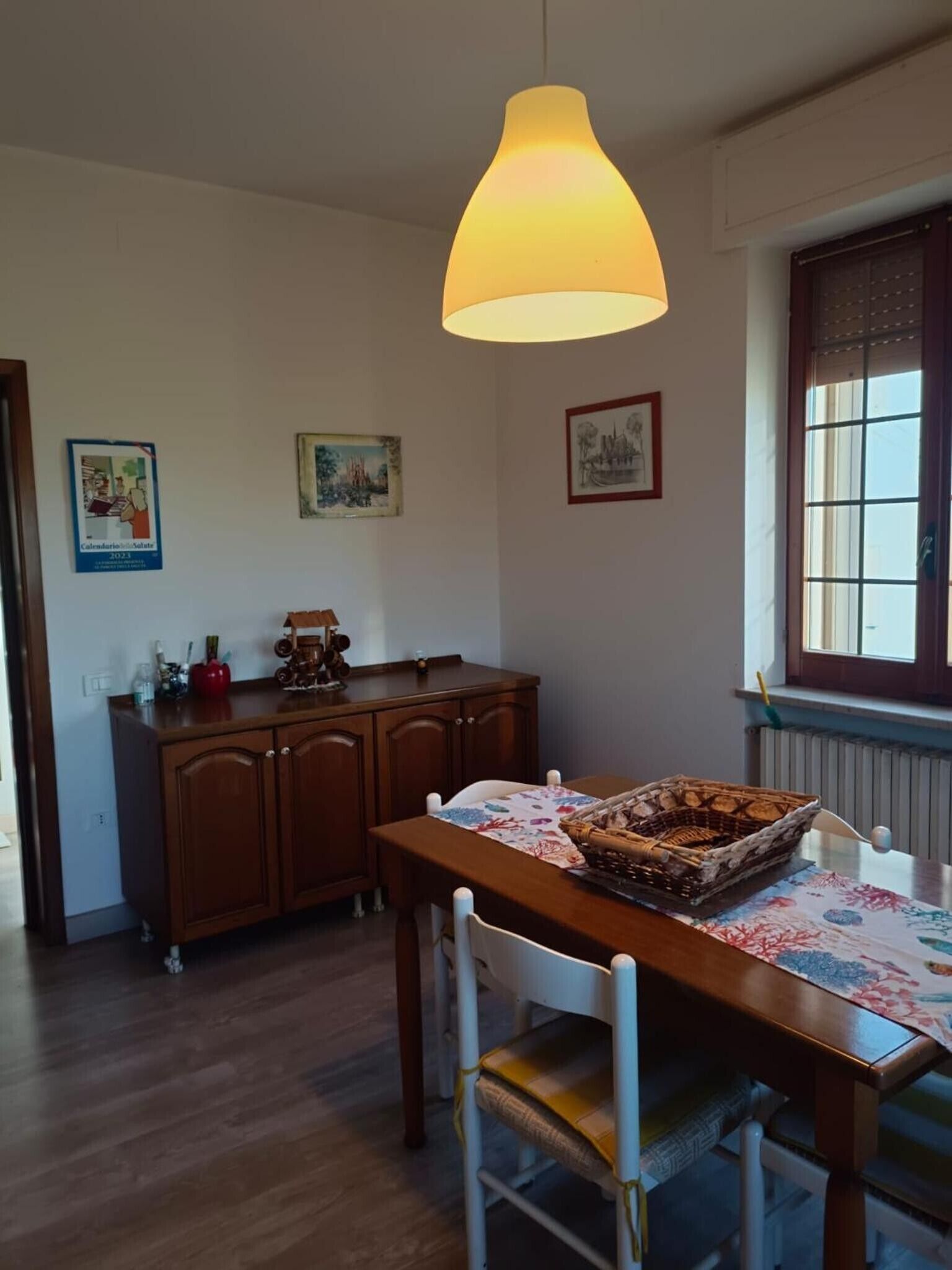 A dining area with a wooden table, chairs, a hanging light fixture, and a window with blinds.