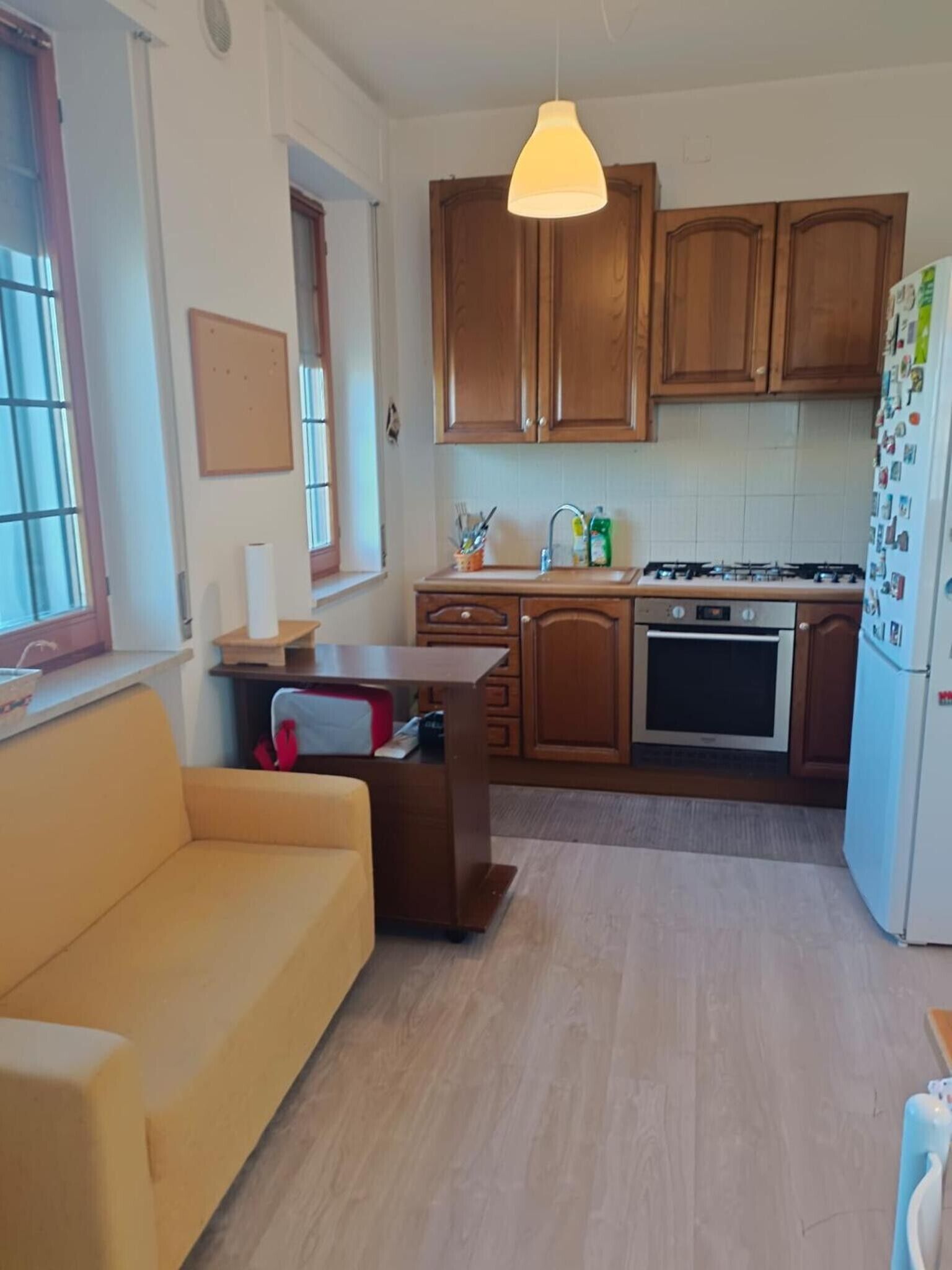 Cozinha compacta com armários de madeira, fogão a gás, geladeira e sofá amarelo.
