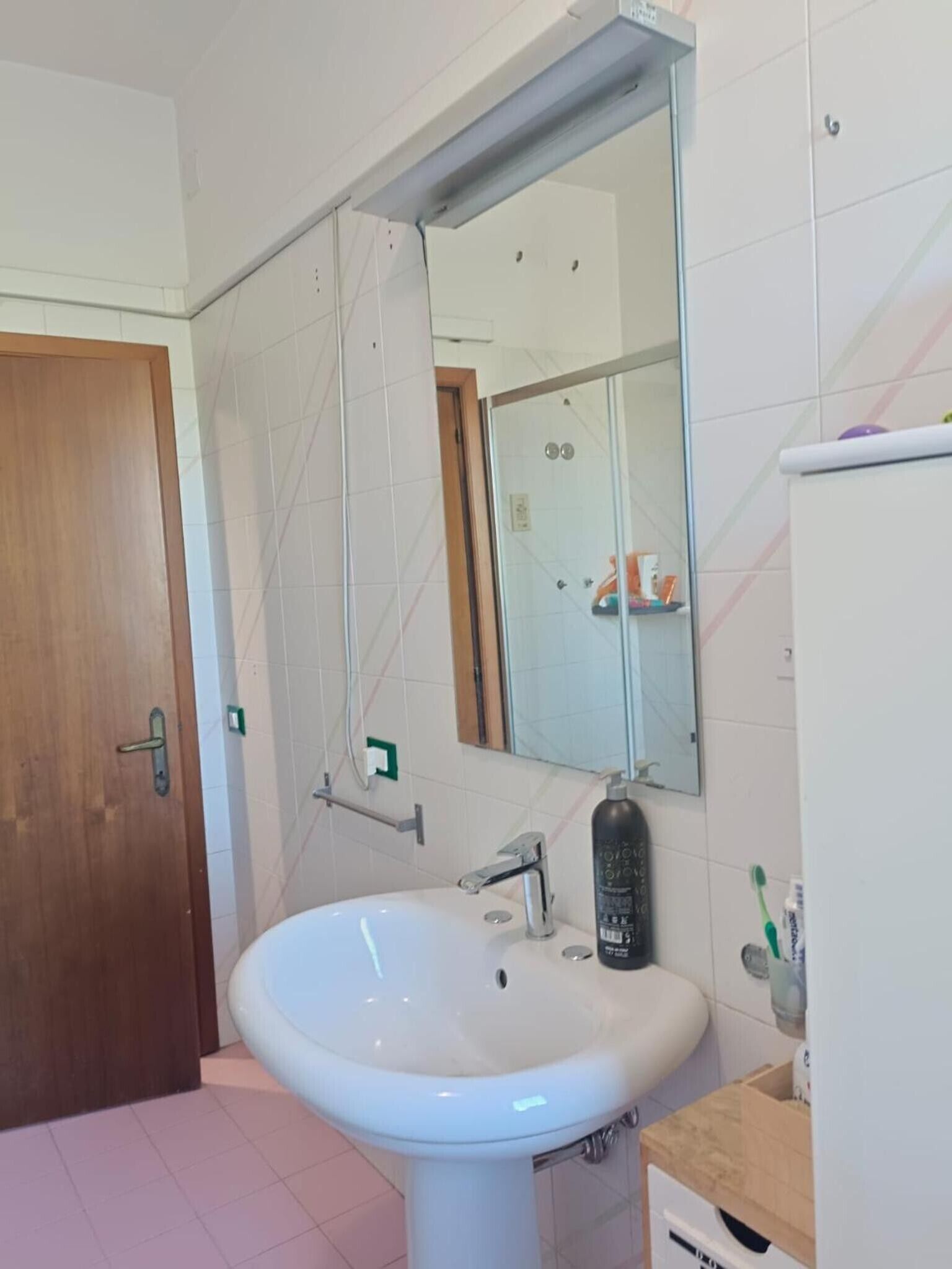 Ein Badezimmer mit weißem Waschbecken, einem Spiegel, einer Dusche und einer Holztür.