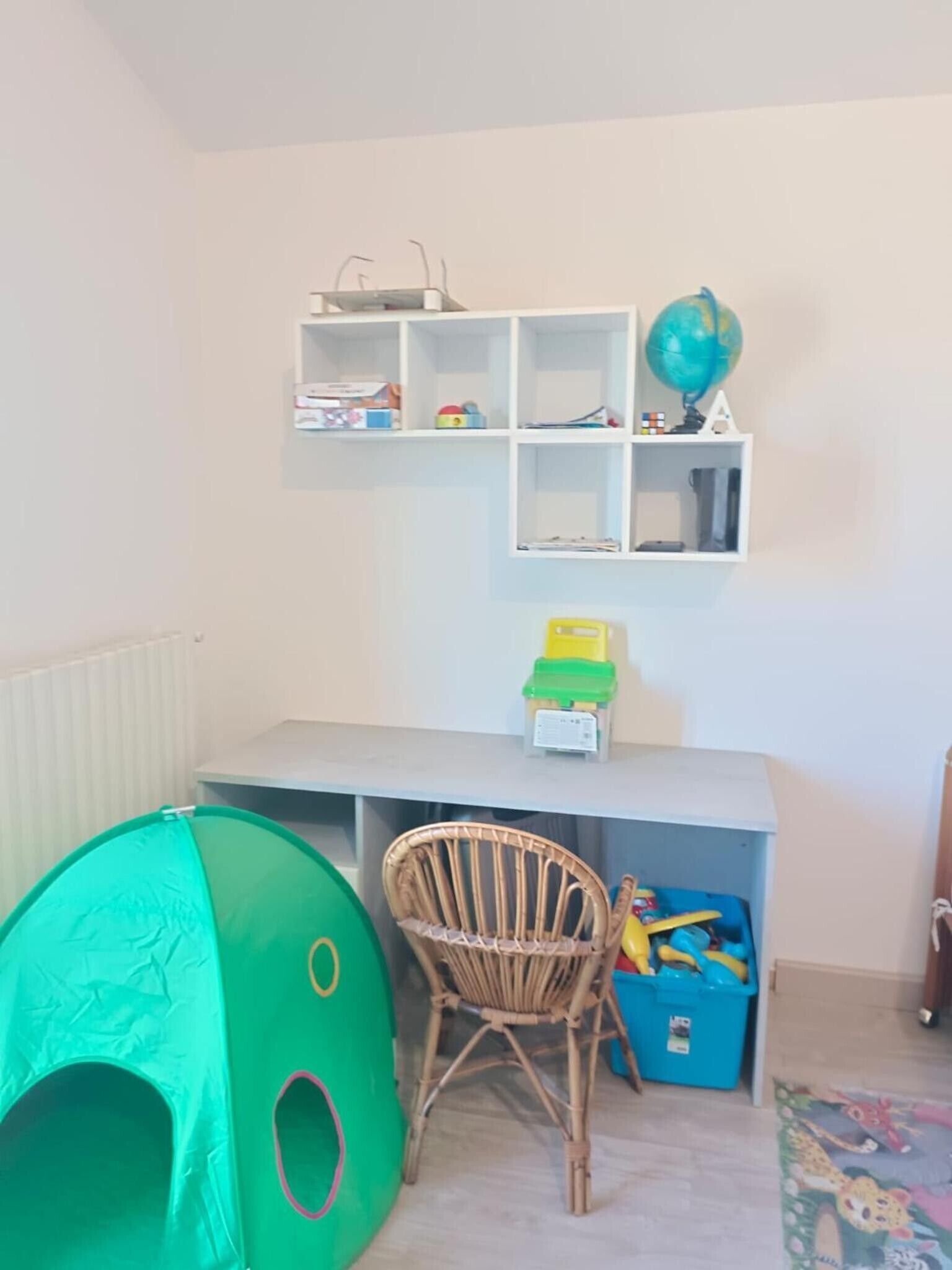 Un área de juegos infantil con una carpa verde, una silla de mimbre y estantes con juguetes y un globo terráqueo.