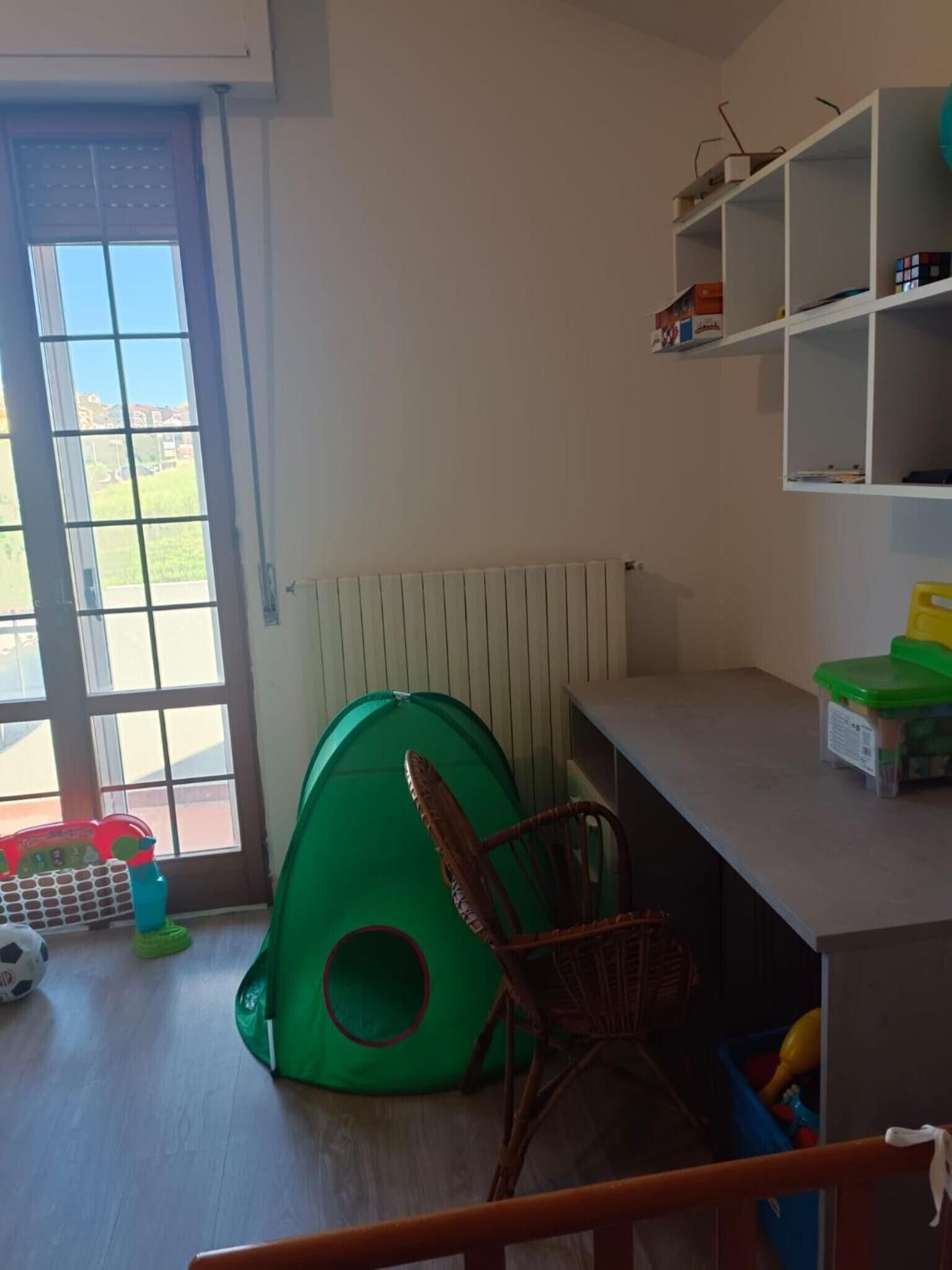 Una habitación con un ventanal grande, una tienda de juegos verde, una silla de mimbre y un radiador blanco.