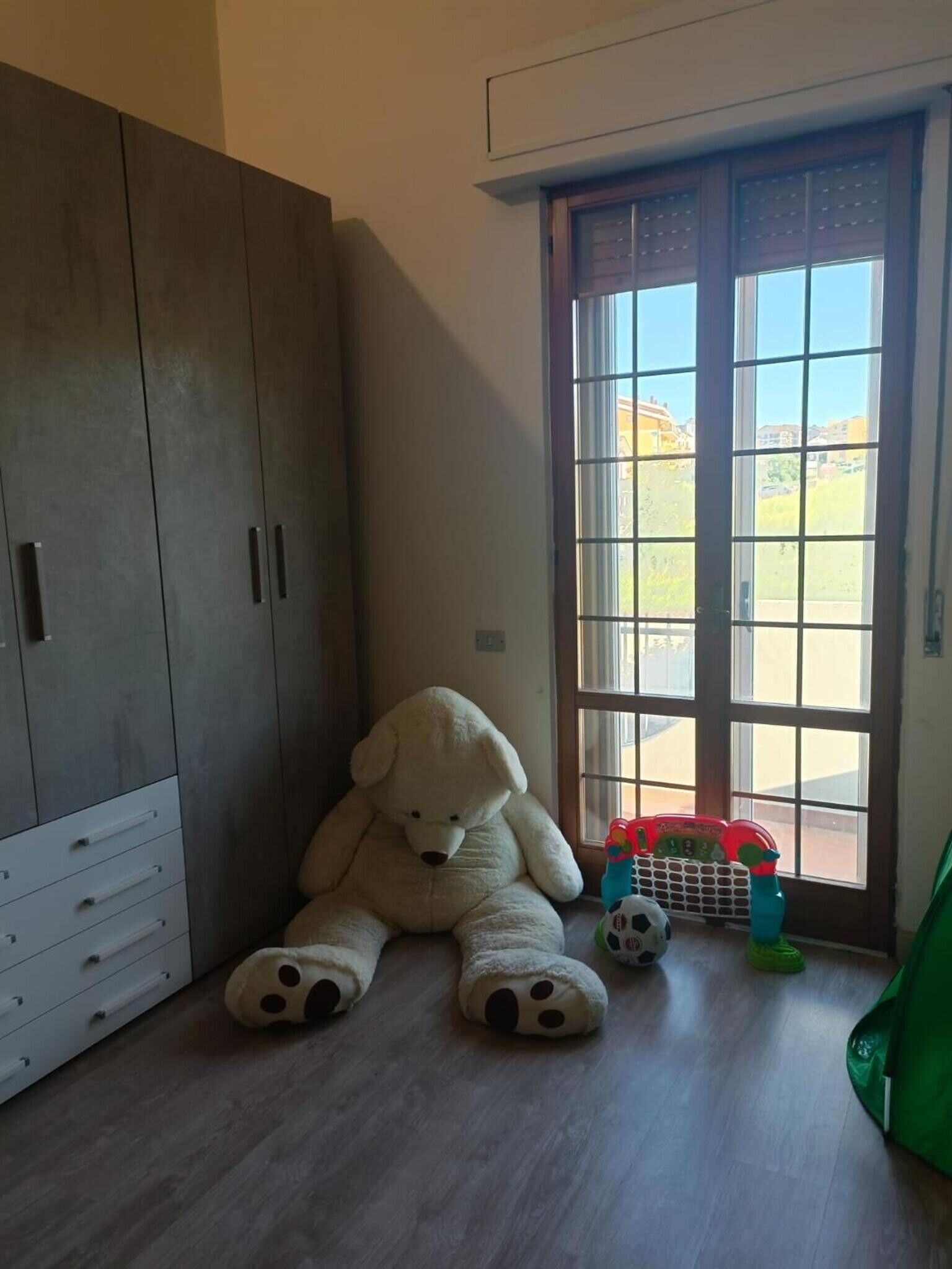 A room with a large wardrobe, a teddy bear, and a window with a view of buildings.