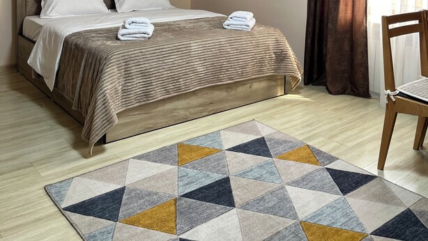 Una cama bien hecha con almohadas y una manta, una silla de madera, un tapete estampado y una ventana con cortinas.