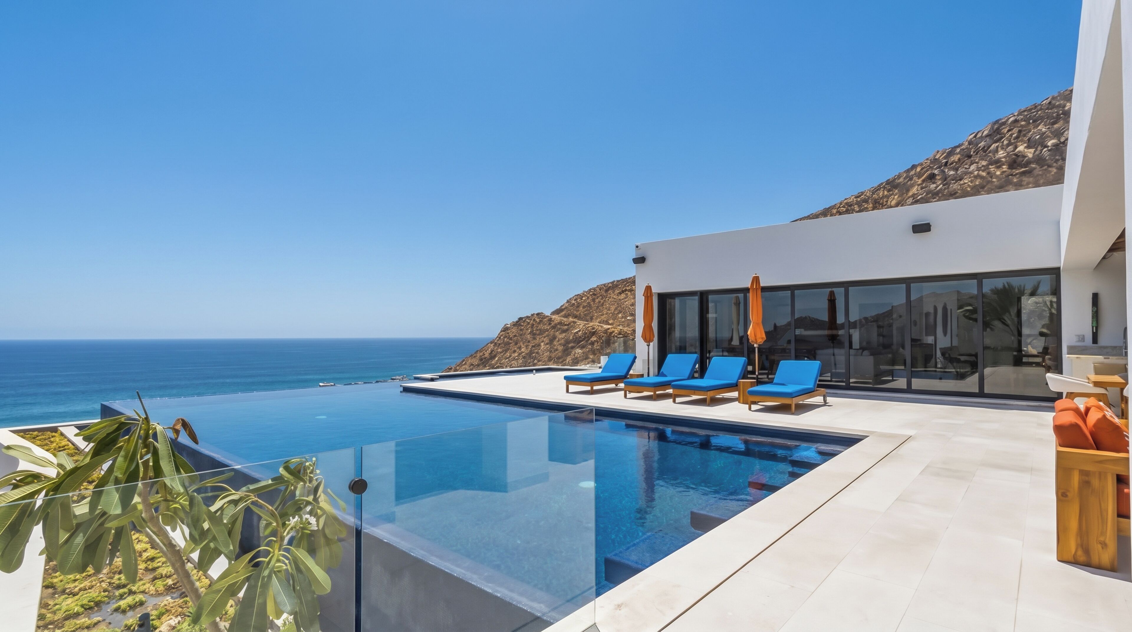 A modern infinity pool with clear blue water, adjacent to a white building with large windows, overlooking a rocky coastline and the ocean.