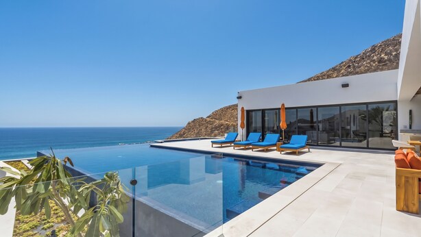 Ein moderner Infinity-Pool mit klarem, blauem Wasser, neben einem weißen Gebäude mit großen Fenstern, mit Blick auf eine felsige Küstenlinie und den Ozean.