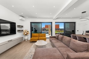 Ein modernes Wohnzimmer mit großem Flachbildfernseher, einer Couch, einem Couchtisch und einem Essbereich mit Tisch und Stühlen.