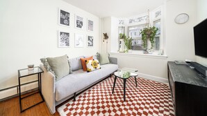 Un salon comprenant un canapé, une table basse, un meuble pour la télévision, un tapis à motifs et une fenêtre avec des plantes.