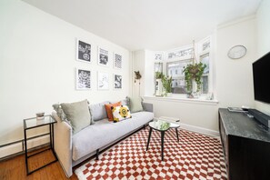 Ein Wohnzimmer mit Sofa, Couchtisch, TV-Schrank, einem gemusterten Teppich und einem Fenster mit Pflanzen.
