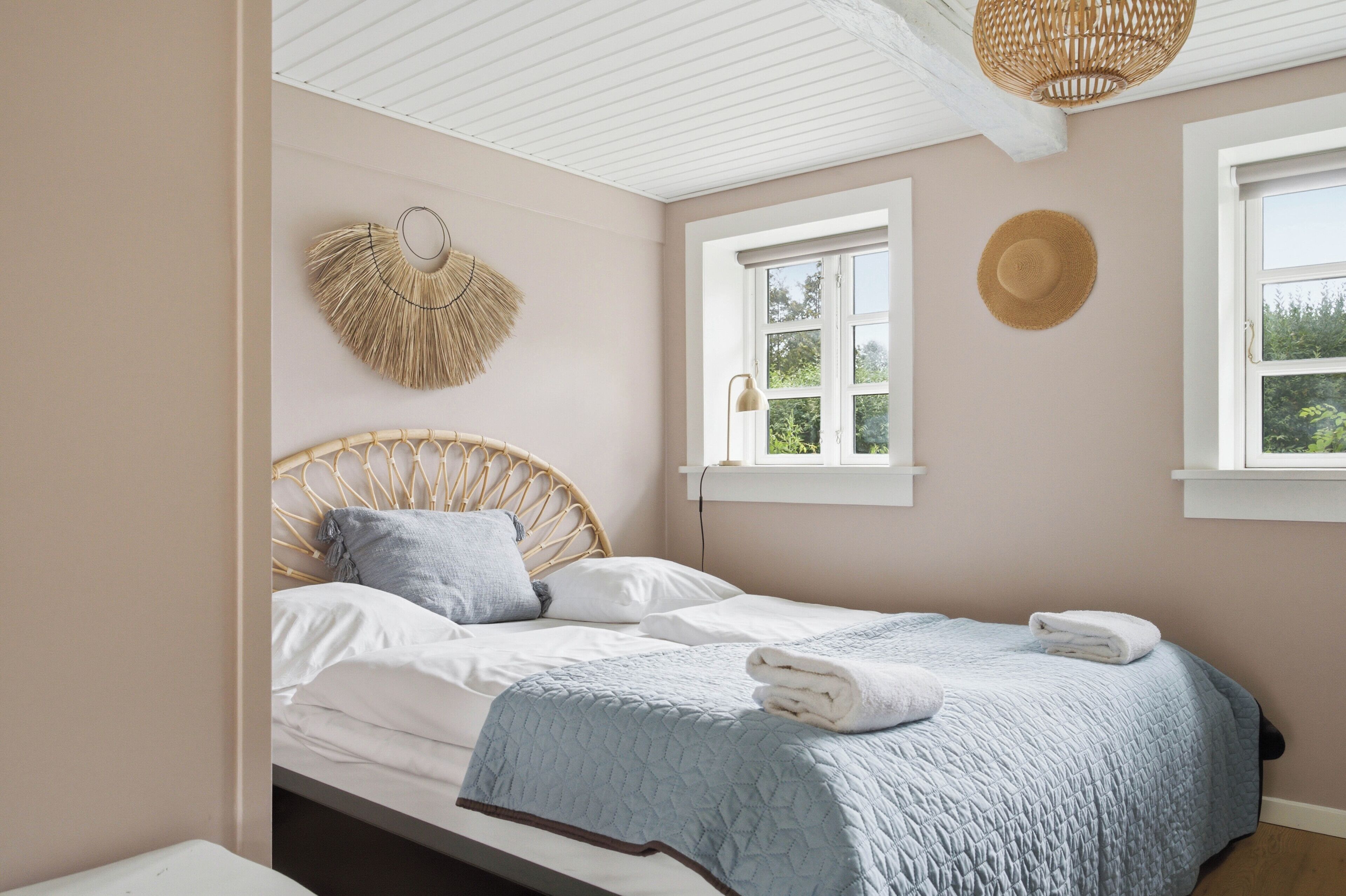 A bedroom with a bed, two windows, a woven wall decoration, and a straw hat on the wall.