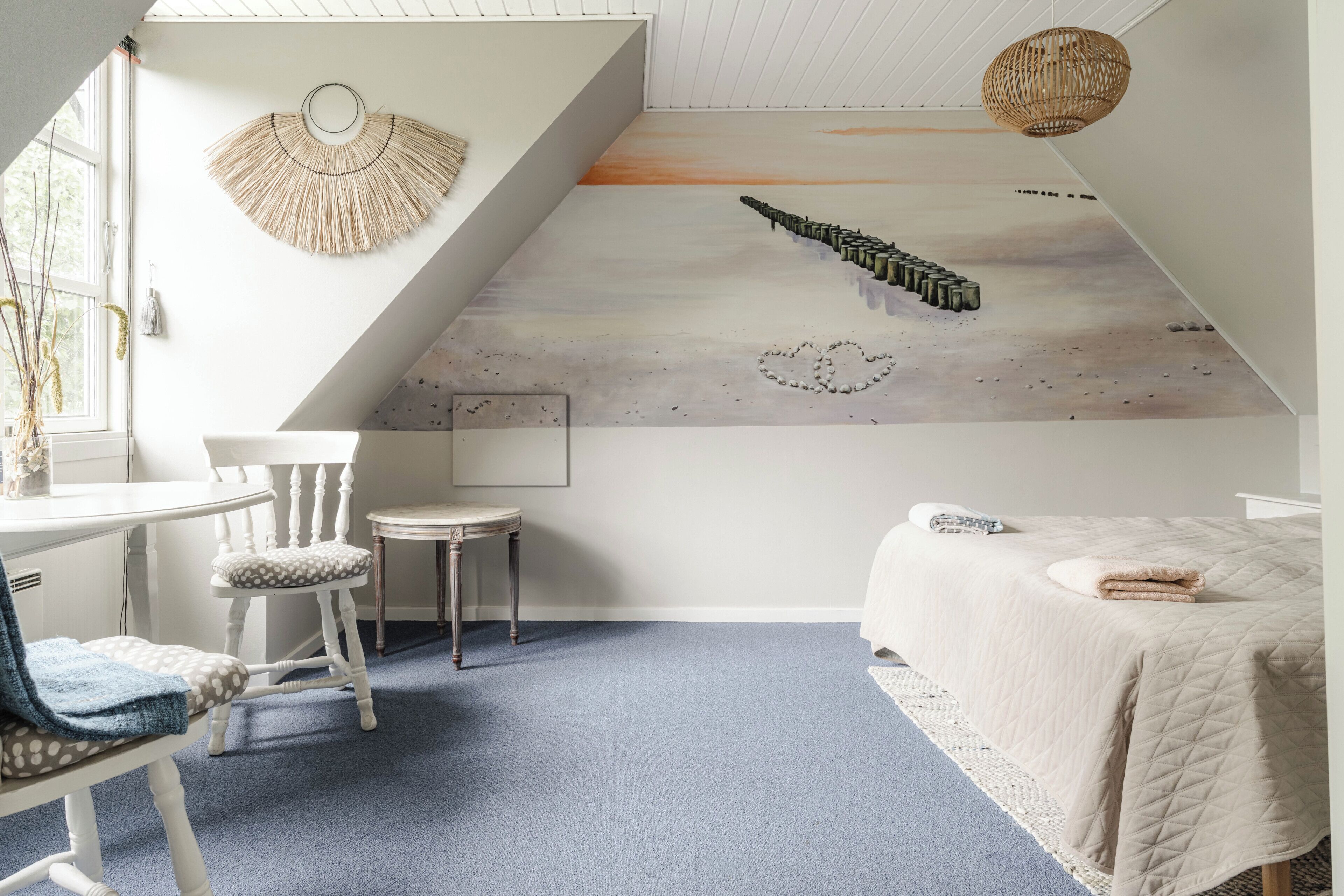 A bedroom with a sloped ceiling, a bed with a quilted cover, a chair, a small table, and a wall mural.