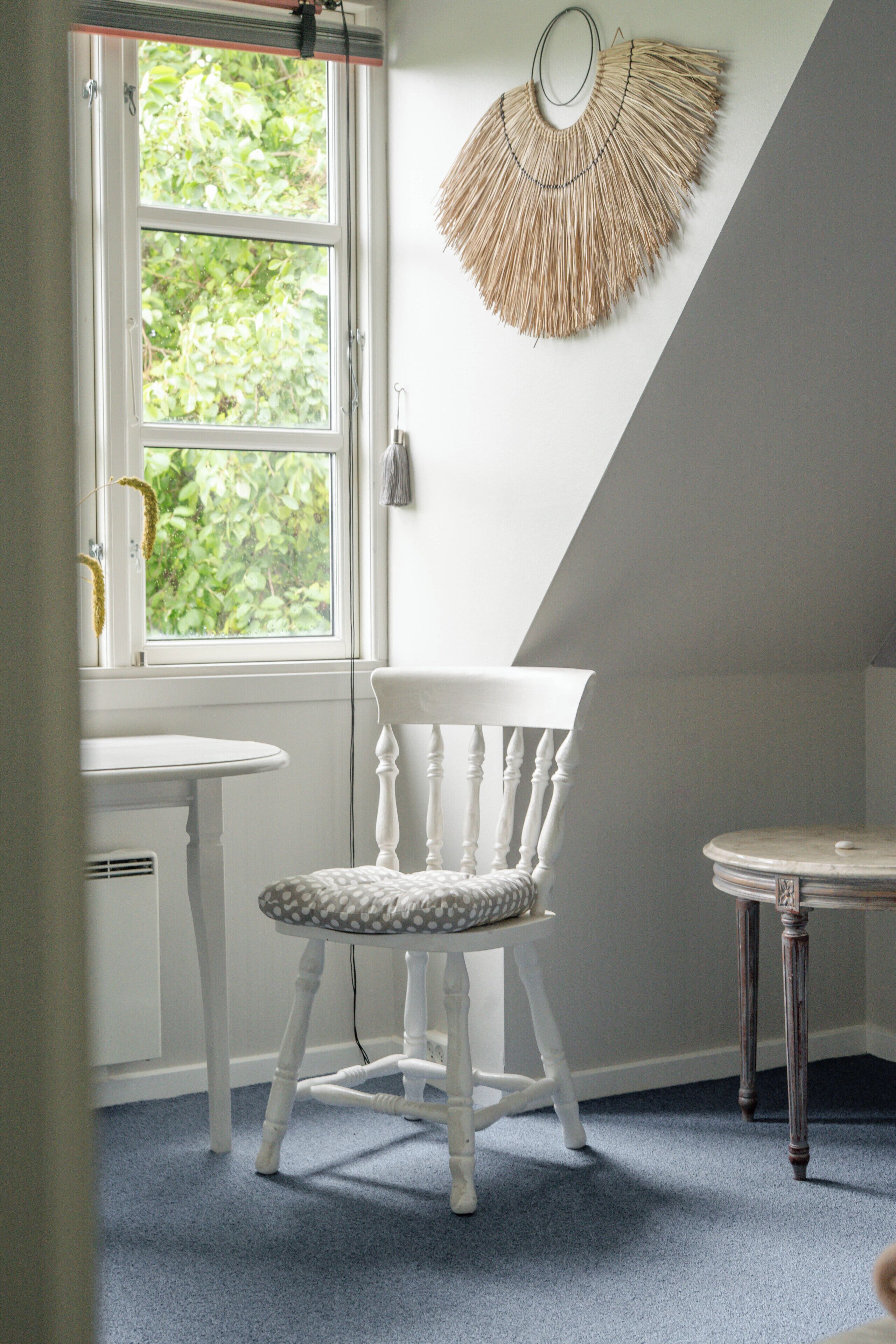 A room with a white chair, a small round table, a window with a view of greenery, and a wall-mounted decorative item.