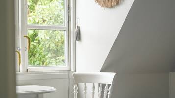 A room with a white chair, a small round table, a window with a view of greenery, and a wall-mounted decorative item.