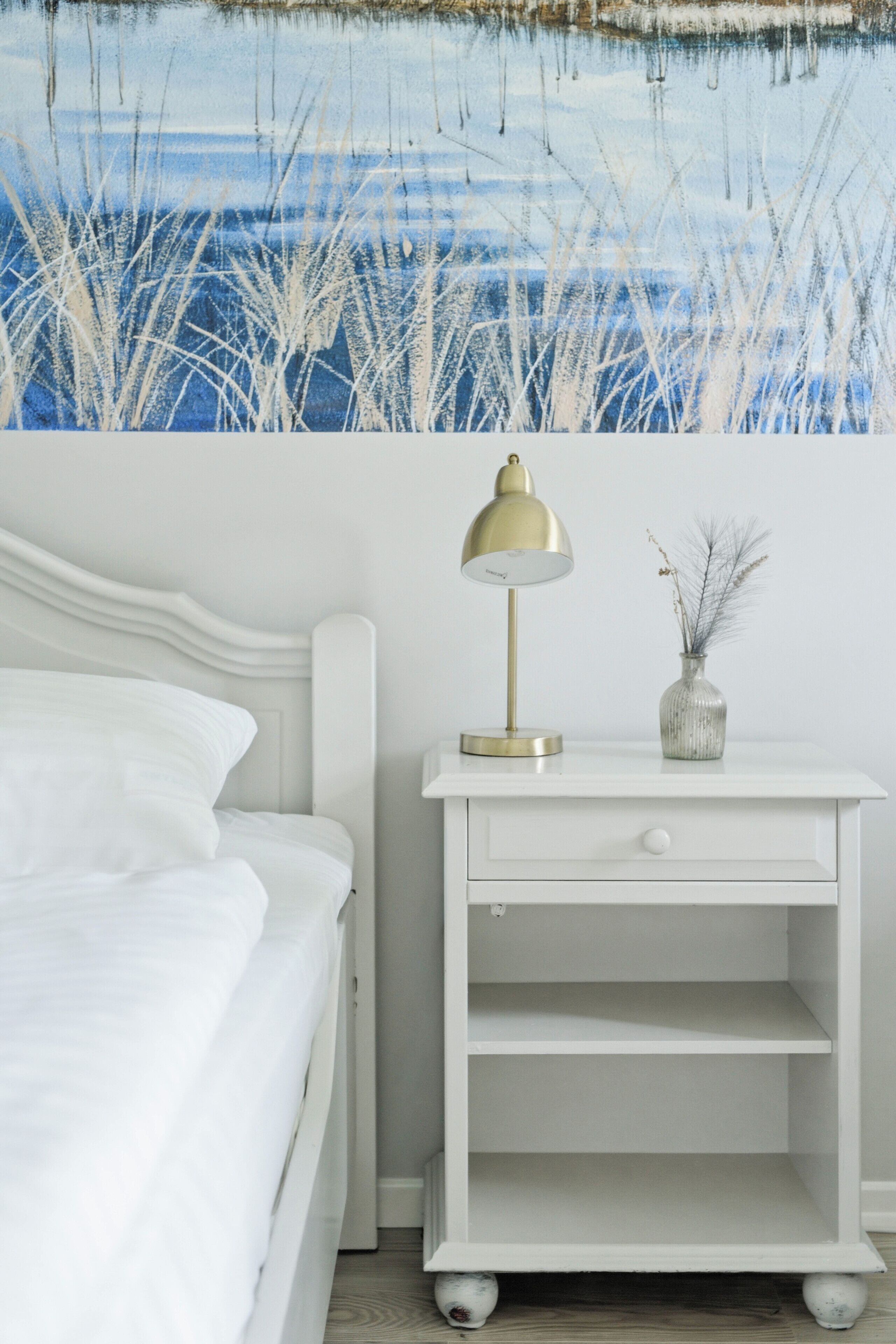 A bedroom with a white bed, a bedside table with a lamp, and a wall painting.