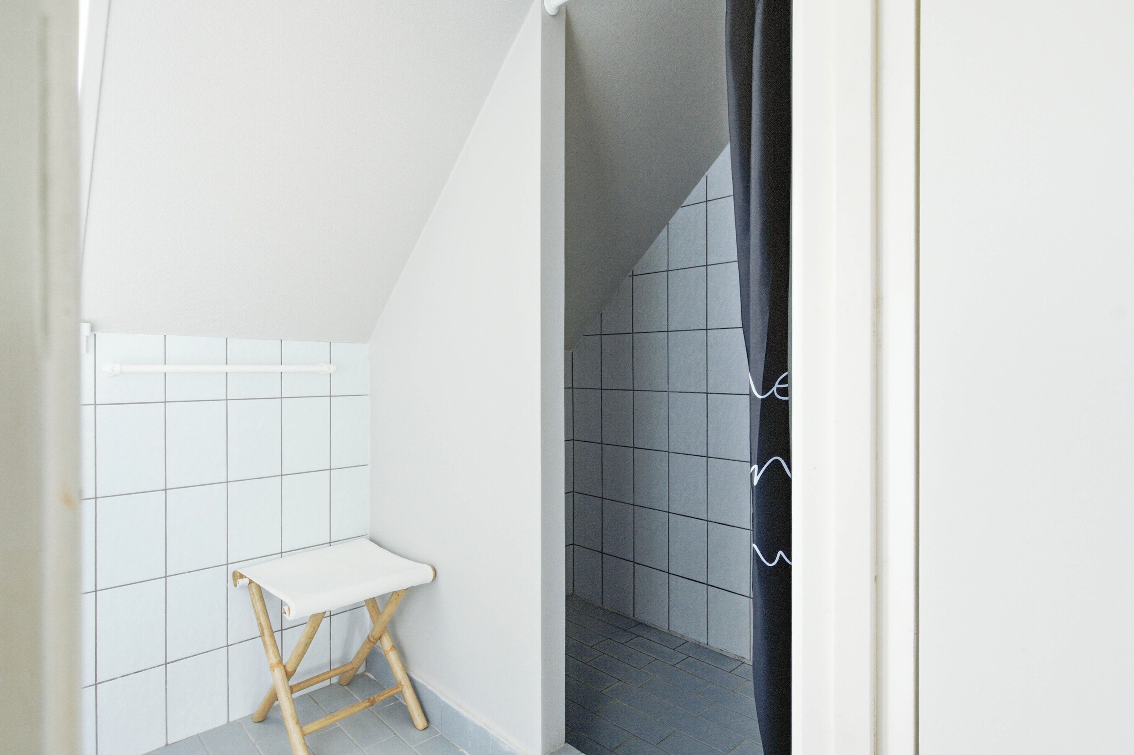A small, narrow bathroom with white and gray tiles, a white bench, and a black shower curtain.