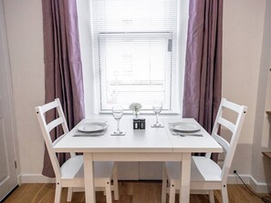 A dining table with white chairs, a window with blinds, and a vase with flowers.