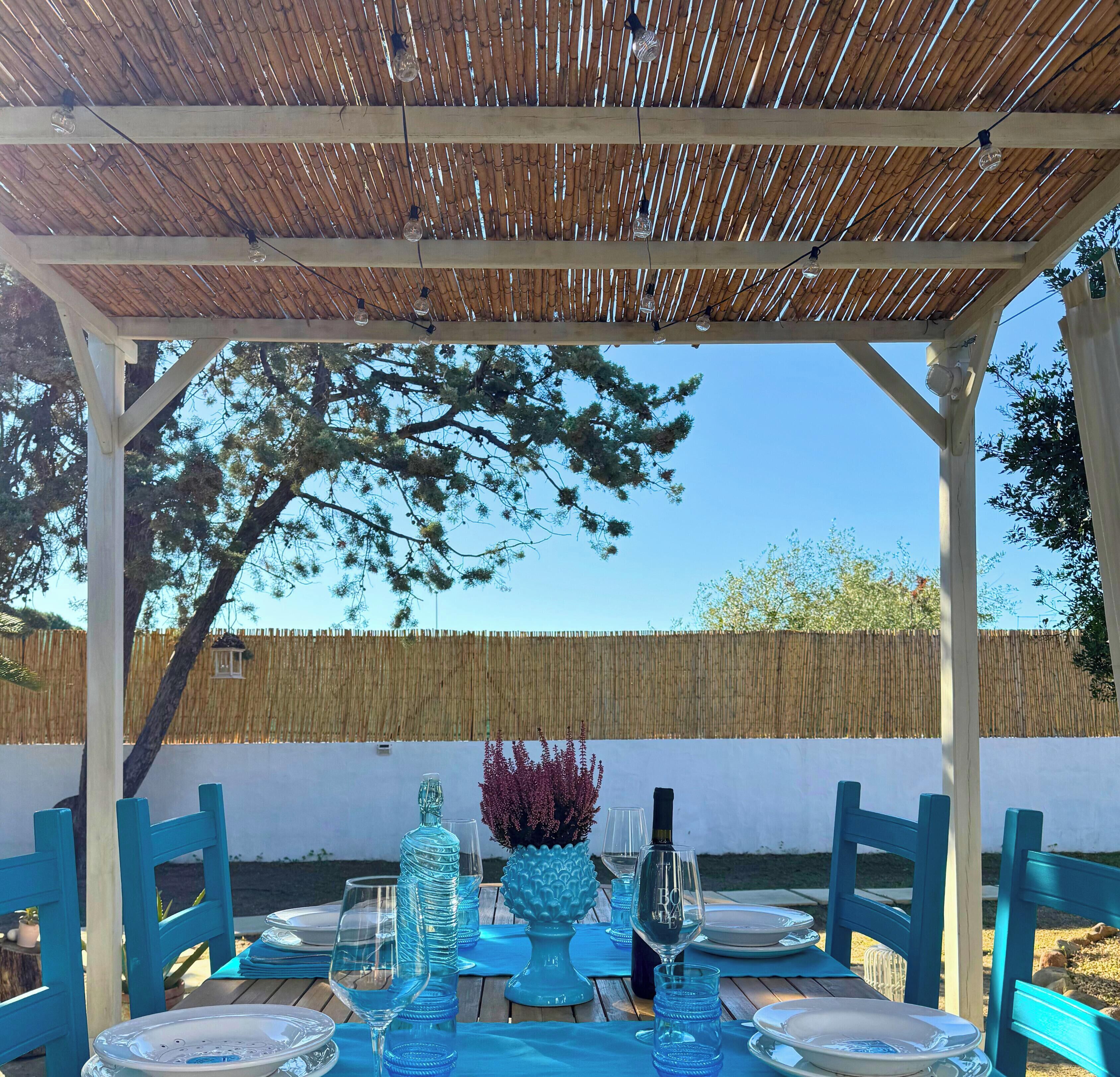 An outdoor patio with a bamboo roof, a table set with blue glassware, and a decorative vase.