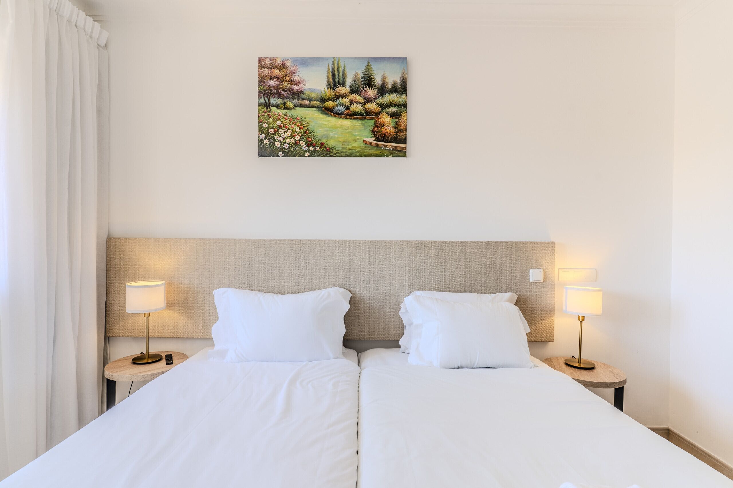 Un lit double avec du linge de lit blanc, deux tables de chevet avec des lampes, un tableau représentant un paysage et une tête de lit.