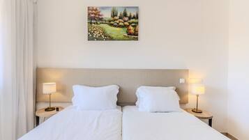 Un lit double avec du linge de lit blanc, deux tables de chevet avec des lampes, un tableau représentant un paysage et une tête de lit.