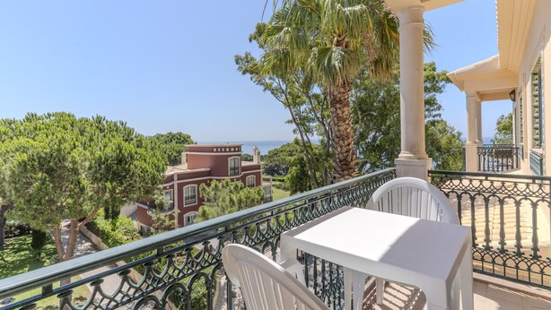 Un balcón con una mesa y sillas blancas, con vistas a una zona costera con árboles y un edificio.