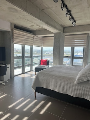 Ein Schlafzimmer mit einem großen Bett, einem Schreibtisch und einem Stuhl, mit Blick auf eine Stadtkulisse durch große Fenster.