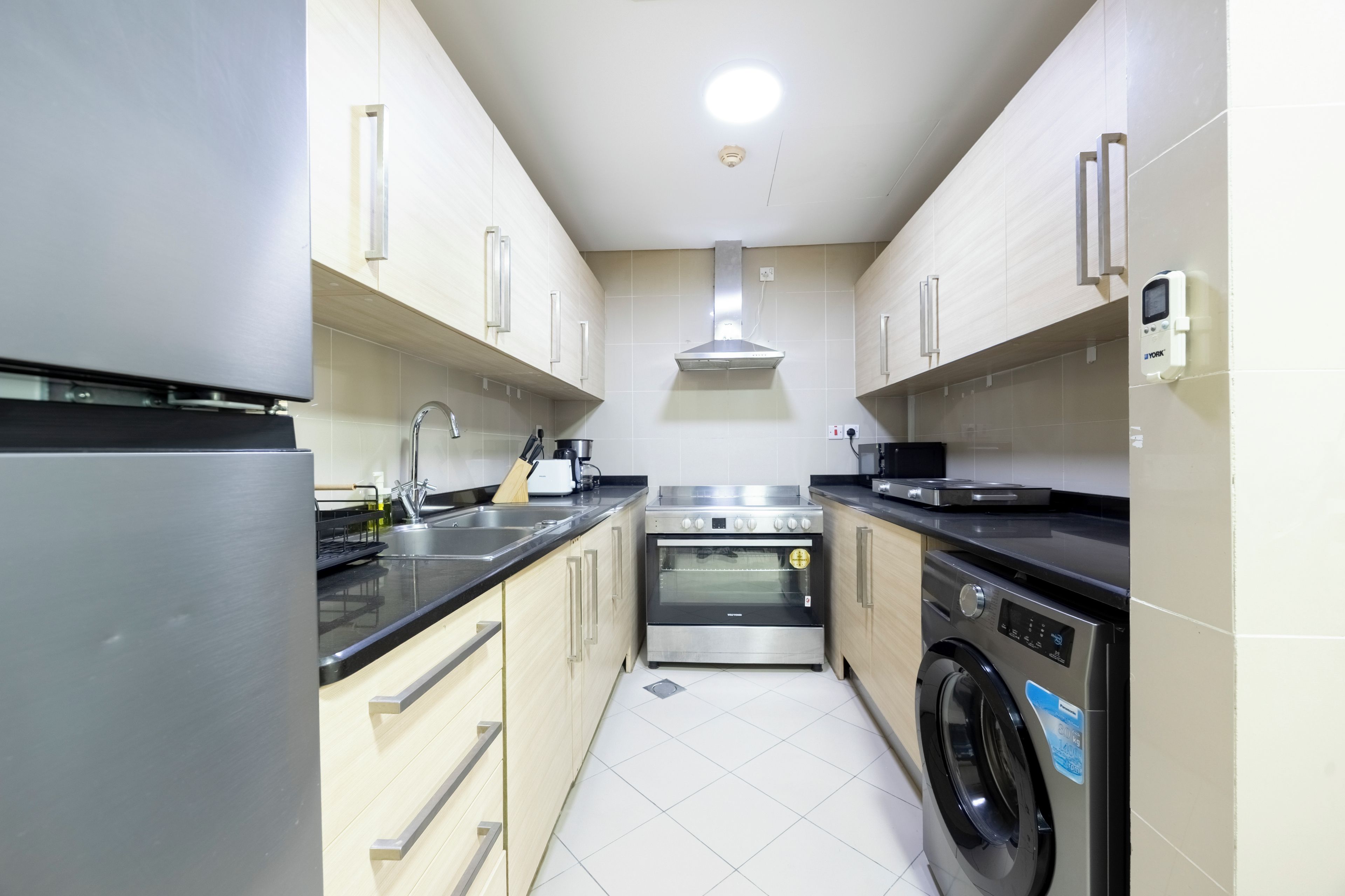 A compact kitchen with a stainless steel refrigerator, a built-in oven, a washing machine, and a sink with modern faucets.
