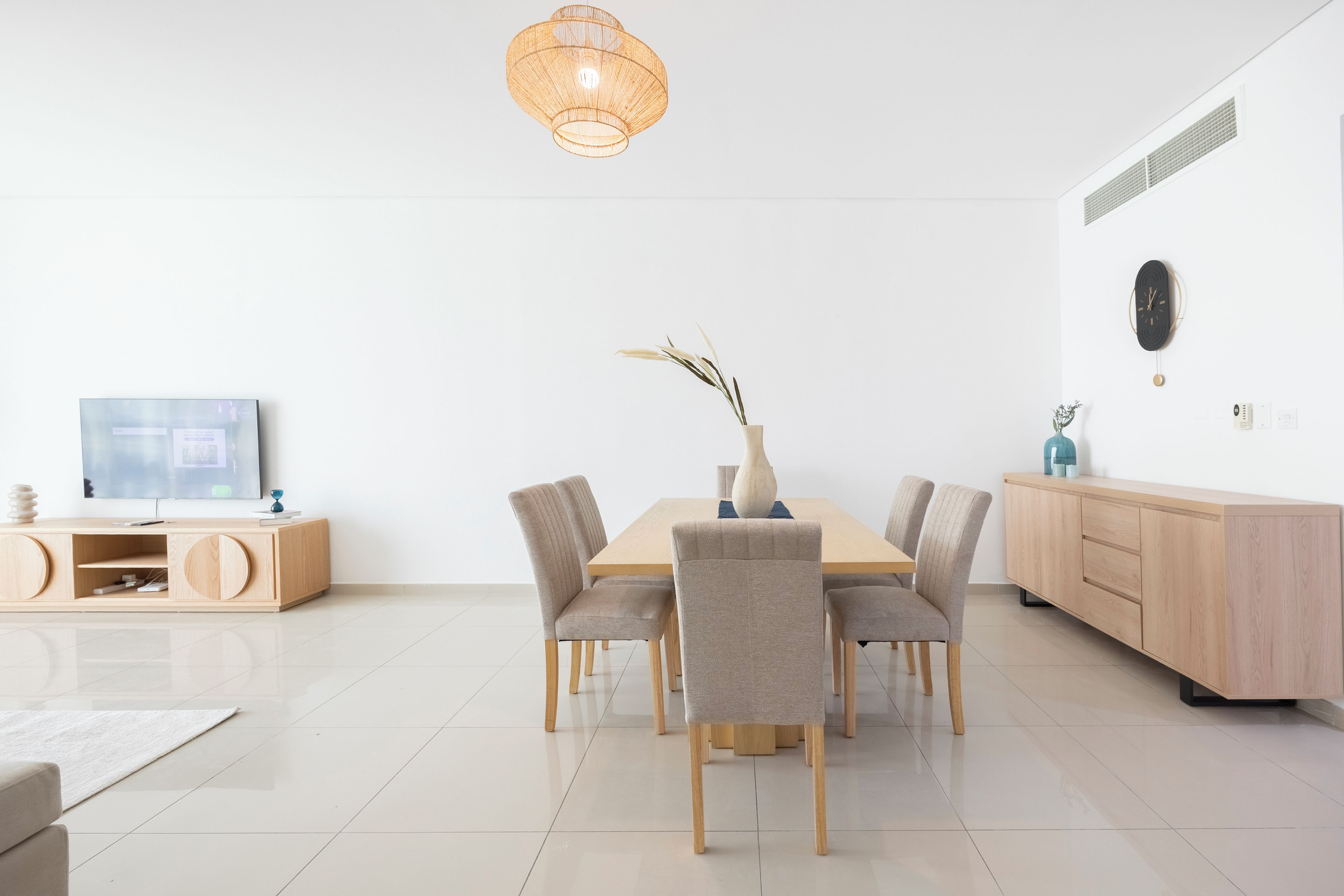 A modern dining area with a wooden table and chairs, a vase with branches, a wall-mounted clock, and a television on a wooden console.