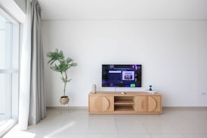 Uma sala de estar moderna com televisão de tela plana fixada na parede, um móvel de madeira embaixo, um vaso de planta e uma janela grande com cortinas.