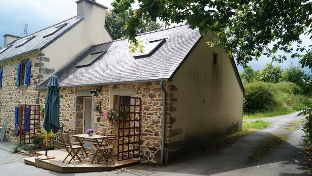 A stone building with a sloped roof, a small porch with a table and chairs, and a window with blue shutters.