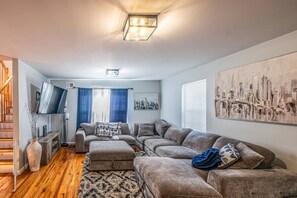A living room with a sofa set, a flat-screen TV, a cityscape painting, and a staircase.