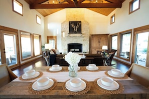 A dining area with a long wooden table set for six, a stone fireplace, and large windows. - The Ranch House (Hondo)